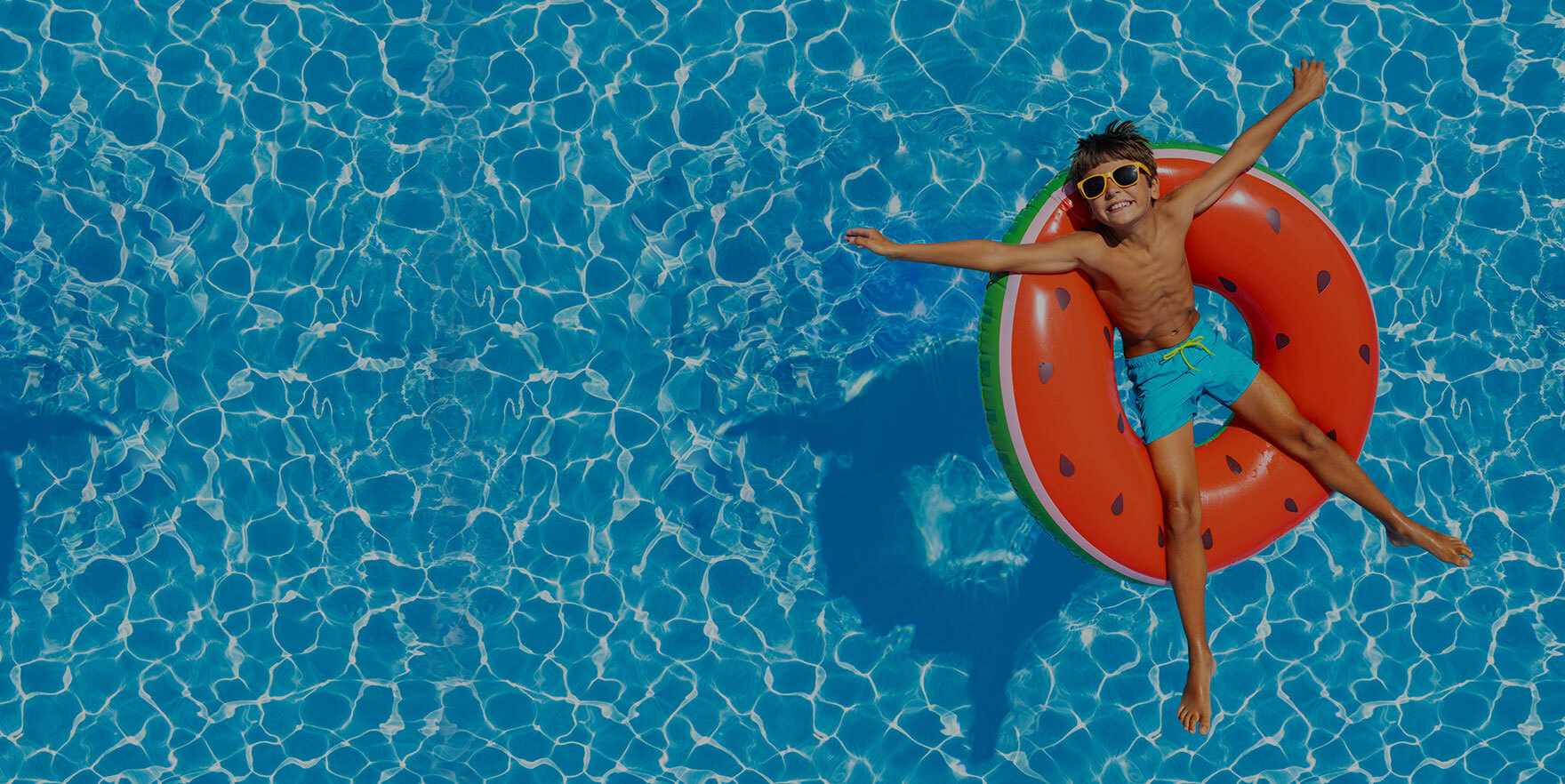 Smiling boy in sunglasses floating on red watermelon pool ring in clear blue water on sunny day